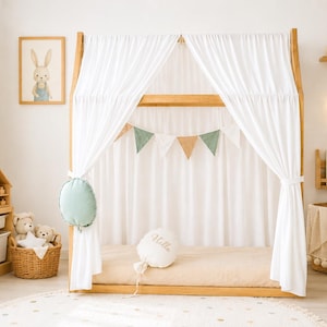 May include: A wooden house-shaped floor bed frame with white fabric curtains, a beige mattress, and a decorative bunting banner. The bed features a round sage green pillow and a white balloon-shaped pillow with the word Hello embroidered on it.