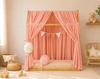 May include: A wooden house-shaped floor bed frame draped with soft coral-colored fabric curtains. A white pennant banner hangs inside. Two balloon-shaped pillows, one white with the text Hello and one coral, are placed on the bed frame.