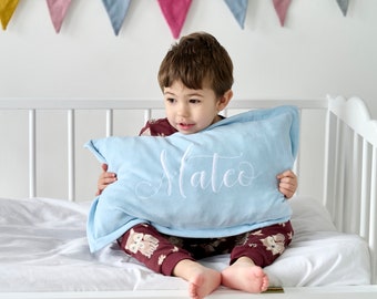 Könnte beinhalten: Ein kleiner Junge sitzt auf einem weißen Babybett und hält ein hellblaues Kissen mit dem Namen "Mateo" in weiß gestickt.