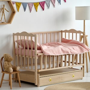 May include: A light wood cot with a pink duvet and pillow. The cot has a drawer with gold-coloured knobs. A small wooden chair with a teddy bear sits nearby. A wooden floor lamp and a decorative wall shelf are also in the room.