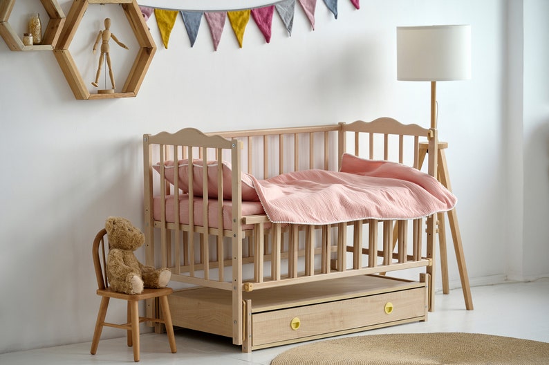 May include: A light wood cot with a pink duvet and pillow. The cot has a drawer with gold-coloured knobs. A small wooden chair with a teddy bear sits nearby. A wooden floor lamp and a decorative wall shelf are also in the room.