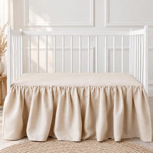 Puede incluir: Una cuna blanca con una sábana bajera de color beige sólido y un volante de cama beige a juego con un diseño fruncido que cuelga para cubrir la base del marco de la cuna, creando una apariencia limpia y neutral.