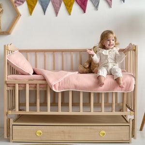 May include: A light wood cot with a pink blanket and pillow. A small child sits on the bed, smiling. The cot has a drawer with yellow knobs. A wooden chair and a floor lamp are nearby.