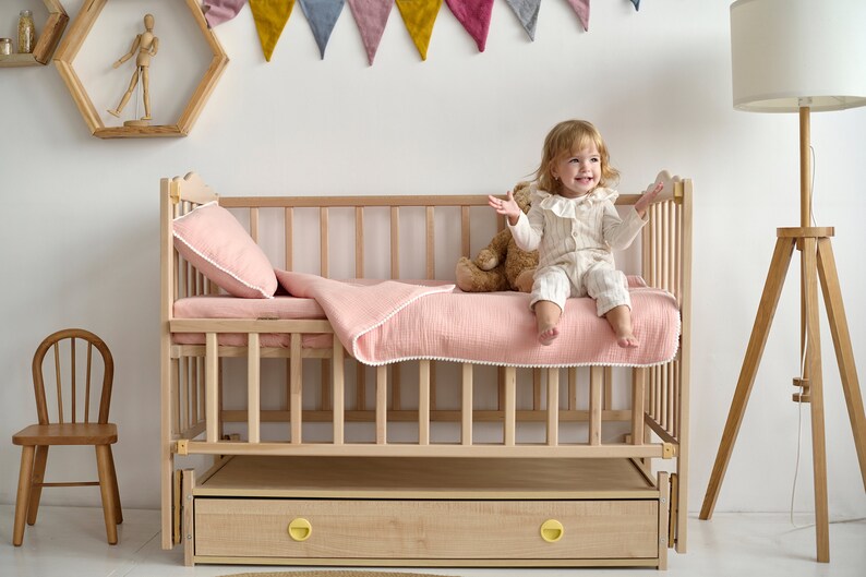 May include: A light wood cot with a pink blanket and pillow. A small child sits on the bed, smiling. The cot has a drawer with yellow knobs. A wooden chair and a floor lamp are nearby.
