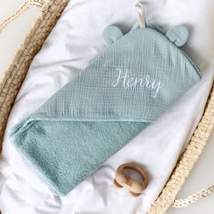 Puede incluir: Una toalla con capucha de color azul claro con el nombre "Henry" bordado en blanco en la parte delantera. La toalla est&aacute; doblada y colocada en una cesta tejida.