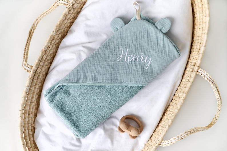Puede incluir: Una toalla con capucha de color azul claro con el nombre "Henry" bordado en blanco en la parte delantera. La toalla est&aacute; doblada y colocada en una cesta tejida.
