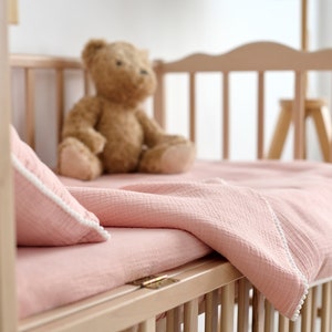 May include: A light-coloured wooden cot with a pink blanket, pillow, and a teddy bear. The bedding has white trim. The cot is empty, ready for a baby.
