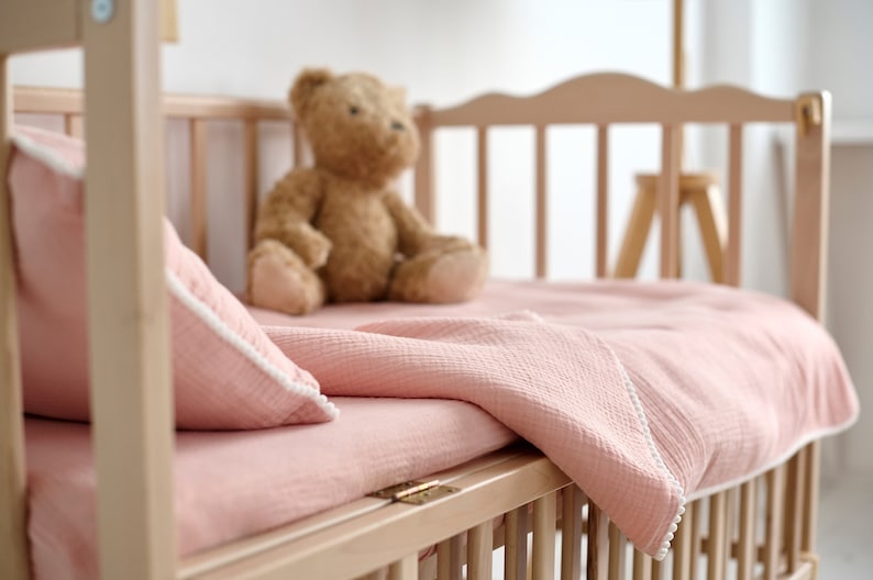May include: A light-coloured wooden cot with a pink blanket, pillow, and a teddy bear. The bedding has white trim. The cot is empty, ready for a baby.