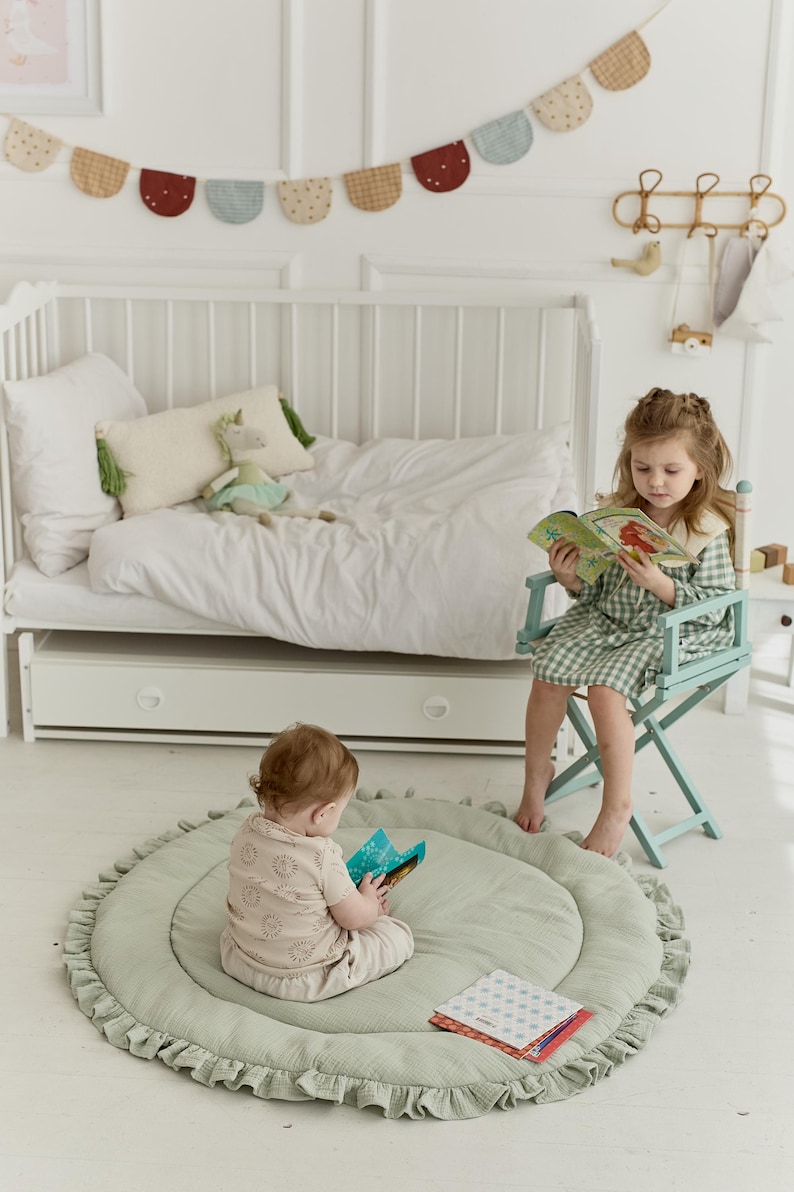 May include: A round, sage green quilted play mat with a ruffled edge, featuring a soft fabric texture. The mat is placed on a white floor, providing a comfortable surface for a child sitting and reading a book, with a second book resting nearby.