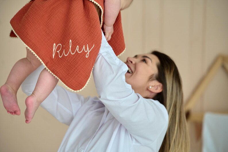 May include: A rust-coloured muslin blanket featuring the name Riley embroidered in white script lettering. The blanket has a textured fabric surface and is finished with a delicate cream-coloured pom-pom trim along the edges.