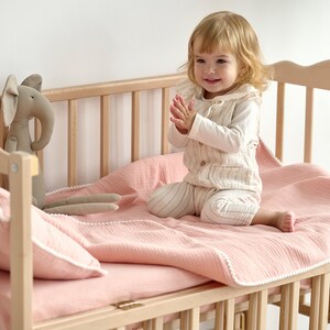 May include: A light wood baby cot with a pink blanket and pillow set. A stuffed elephant toy sits near the pillow. A toddler in a striped outfit sits in the cot, clapping their hands.