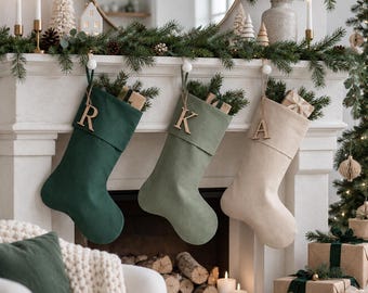 May include: Three Christmas stockings hanging in a row, featuring a dark green, a sage green, and a cream-colored stocking. Each stocking has a wooden letter tag attached to the cuff, displaying the letters R, K, and A respectively.