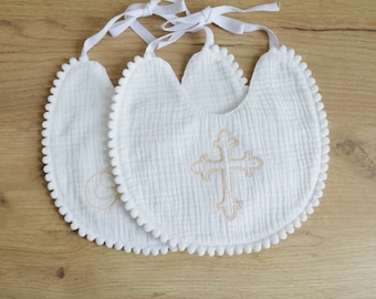 May include: Two white cotton baby bibs with a scalloped edge and pom pom trim. One bib features a gold embroidered cross, the other has a simple design.