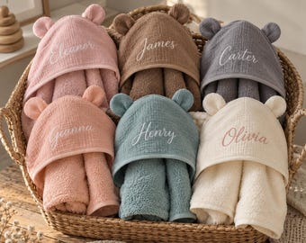 May include: A collection of six hooded baby towels in a woven basket. Each towel features bear ears on the hood and is embroidered with a name: Eleanor, James, Carter, Gianna, Henry, or Olivia. The towels are in pink, brown, gray, peach, blue, and cream.