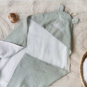May include: A soft, green and white muslin hooded towel with the name "Lucas" embroidered on it. The towel is folded and lying on a beige rug.