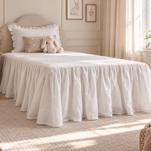 Könnte beinhalten: Eine weiße Tagesdecke mit einem langen, gerafften Volant, der bis zum Boden reicht. Das Bett ist mit einem weißen Kissen mit Rüschen, einem kleinen beigen bestickten Kissen und einem cremefarbenen Stoffhasen dekoriert.