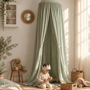 May include: A sage green fabric canopy with a conical top, suspended from the ceiling to create a floor-length draped enclosure. The lightweight, textured material hangs in soft vertical folds, featuring two tie-backs to hold the front panels open.