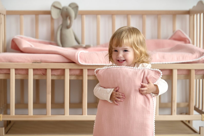 May include: A smiling child holds a pink baby sleep sack with white pom-pom trim. A matching blanket and stuffed elephant are on a wooden cot. The bedding and sleep sack are a soft pink colour.