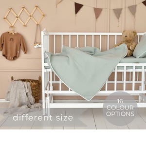 May include: A white crib with a light green linen crib sheet and a stuffed teddy bear. The crib has a white wooden frame and a white mattress. The crib sheet has a white trim. The teddy bear is brown and has a white belly. The crib is in a room with a light pink wall and a white floor. The text "16 Colour Options" and "different size" is visible in the image.