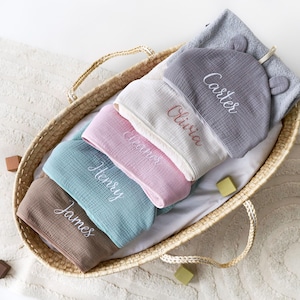 Puede incluir: Una cesta de mimbre llena de cinco toallas con capucha, cada una bordada con un nombre diferente: Carter, Olivia, Amos, Henry y James. Las toallas son de varios colores, incluyendo gris, blanco, rosa, azul y marr&oacute;n.