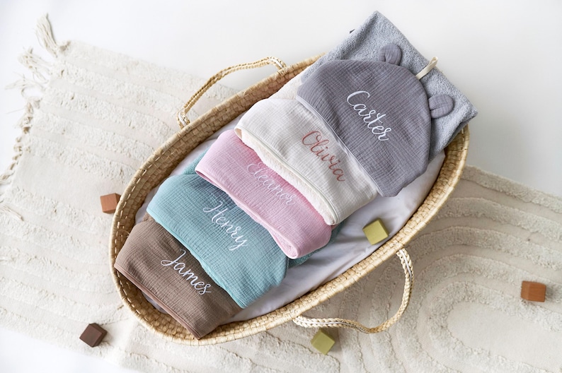 Puede incluir: Una cesta de mimbre llena de cinco toallas con capucha, cada una bordada con un nombre diferente: Carter, Olivia, Amos, Henry y James. Las toallas son de varios colores, incluyendo gris, blanco, rosa, azul y marr&oacute;n.