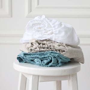 May include: A stack of three linen chair cushions in white, beige, and teal colors. The cushions are folded and stacked on a white stool.