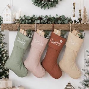 May include: Four fabric Christmas stockings in sage green, dusty pink, terracotta, and beige, each featuring a folded cuff with embroidered names: Daddy, Mommy, Charlie with a paw print, and Eloise. Each stocking contains a small gift box.