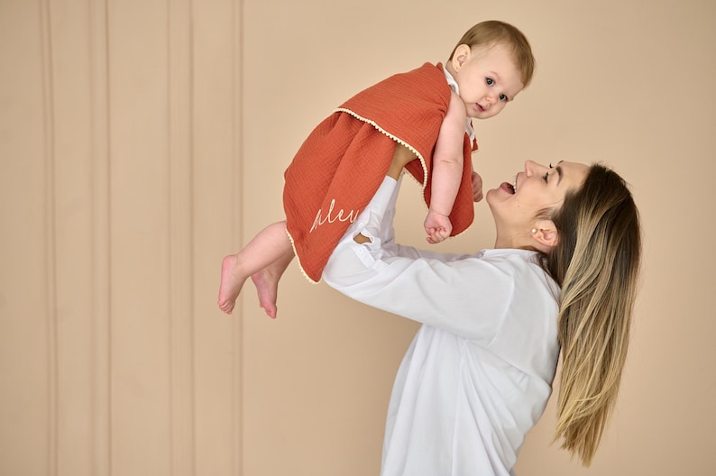 May include: A rust-coloured, textured muslin baby blanket featuring a delicate white pom-pom trim along the edges and cursive white embroidery. The fabric has a soft, crinkled appearance, providing a lightweight and breathable layer for an infant.