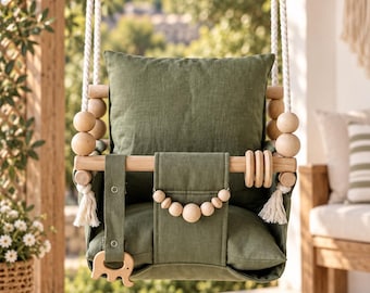May include: A hanging baby swing featuring olive green fabric cushions and natural light-colored wooden beads. The swing is suspended by thick white ropes and includes a wooden front bar with a small wooden elephant-shaped toy attached.
