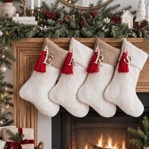 Könnte beinhalten: Vier weiße Plüsch-Weihnachtsstrümpfe mit beigen Bündchen, jeweils mit einem hängenden Holzbuchstaben E, A, R oder S. Jeder Strumpf ist mit einer roten Quaste und einer Holzperlenkette am oberen Bündchen verziert, als Teil der Weihnachtsdekoration.