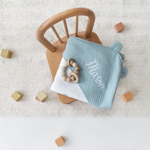 May include: A light blue muslin baby blanket with a white and light blue design. The blanket is personalized with the name "Mason" in white embroidery. A wooden teether is on top of the blanket.