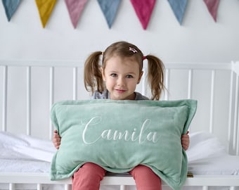 May include: A young girl with pigtails sits in a white crib holding a light green pillow with the name "Camila" embroidered in white.
