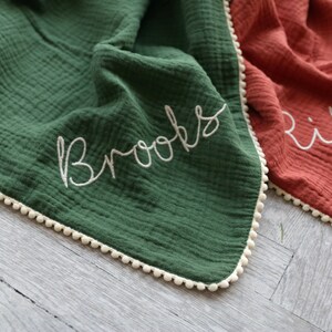 May include: A dark green muslin blanket with a white pom-pom trim and the name Brooks embroidered in white cursive script. Beside it lies a rust-coloured muslin blanket with a white pom-pom trim and the name Riley embroidered in white cursive script.