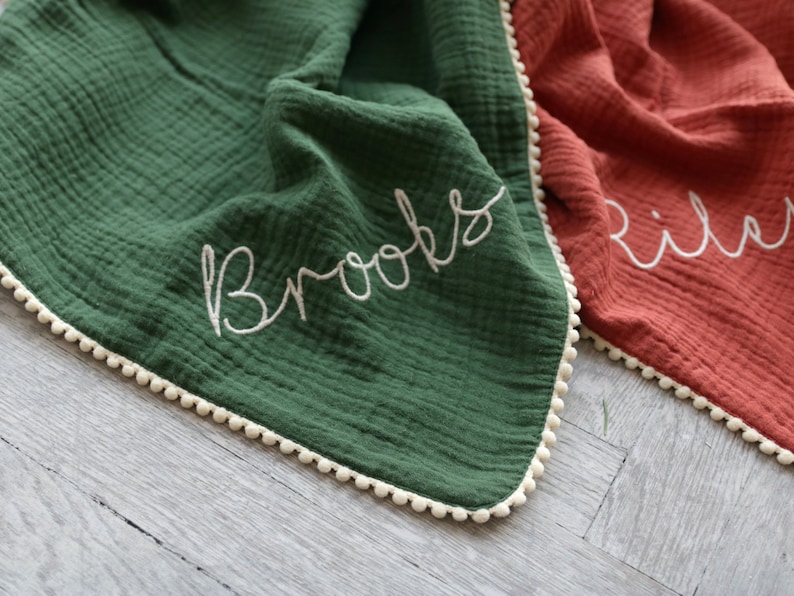 May include: A dark green muslin blanket with a white pom-pom trim and the name Brooks embroidered in white cursive script. Beside it lies a rust-coloured muslin blanket with a white pom-pom trim and the name Riley embroidered in white cursive script.