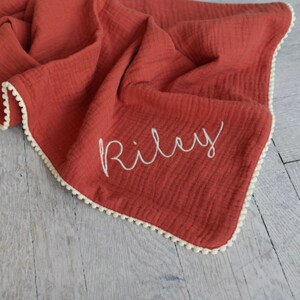 May include: A terracotta-coloured muslin blanket featuring a soft, crinkled texture and a white pom-pom trim along the edges. The name Riley is embroidered in white cursive script on the front of the fabric, creating a distinct contrast.