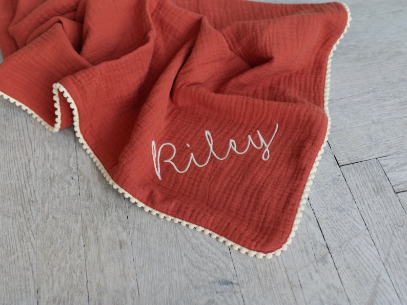 May include: A terracotta-coloured muslin blanket featuring a soft, crinkled texture and a white pom-pom trim along the edges. The name Riley is embroidered in white cursive script on the front of the fabric, creating a distinct contrast.