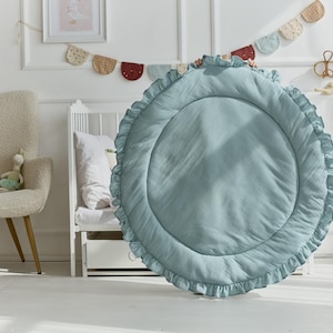 May include: A large, circular, sage green play mat featuring a quilted center and a ruffled border. The mat is made of a soft, textured fabric and is held upright, displaying its round shape and decorative pleated edge detail clearly.