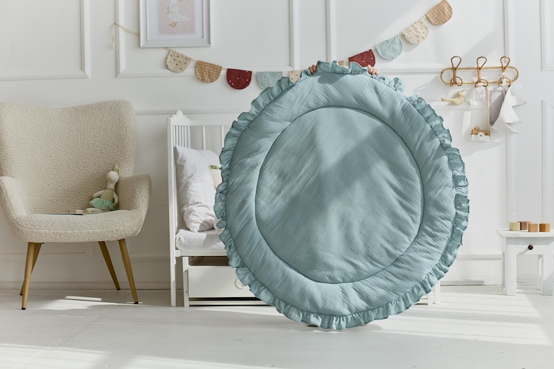 May include: A large, circular, sage green play mat featuring a quilted center and a ruffled border. The mat is made of a soft, textured fabric and is held upright, displaying its round shape and decorative pleated edge detail clearly.