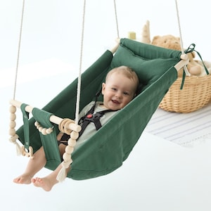 May include: A baby smiles in a dark green canvas swing. The swing is suspended by white rope and has wooden elements and a safety harness. A wicker basket with toys is in the background. The swing is designed for babies.