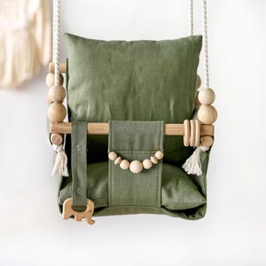 May include: A hanging swing seat made of olive green fabric with a matching back cushion and seat cushion. The frame features natural wood dowels, light-colored wooden beads, white rope suspension, and a wooden elephant-shaped toy attachment.