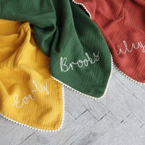 May include: Three muslin blankets in mustard yellow, forest green, and terracotta orange. Each blanket features a white pom-pom trim along the edges and white embroidered names Everly, Brooks, and Riley stitched in a cursive script font.