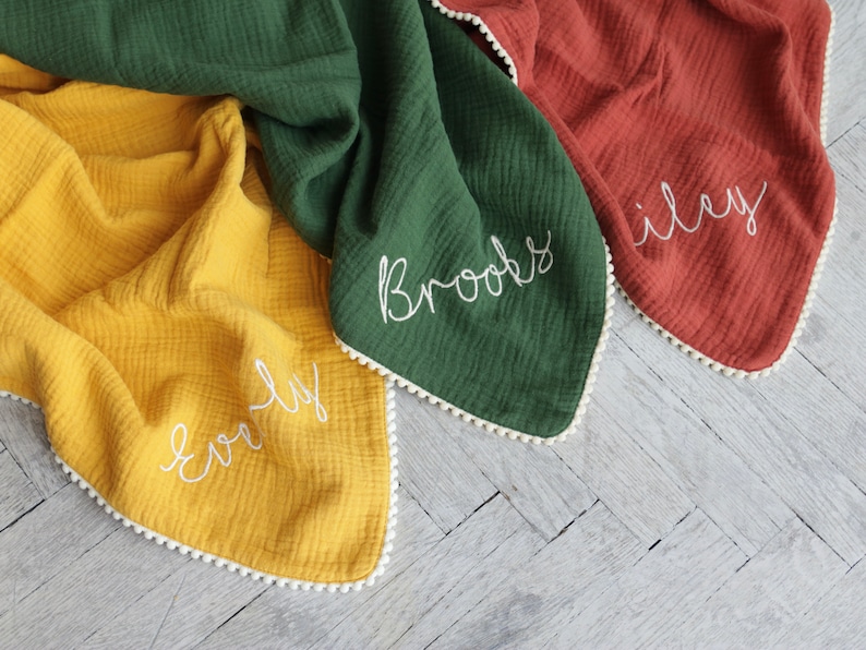 May include: Three muslin blankets in mustard yellow, forest green, and terracotta orange. Each blanket features a white pom-pom trim along the edges and white embroidered names Everly, Brooks, and Riley stitched in a cursive script font.