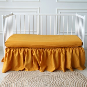 Pode incluir: Uma saia de berço amarela mostarda com uma borda franjada. A saia é feita de um tecido de linho macio e é perfeita para adicionar um toque de elegância a qualquer berçário.