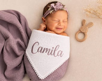 May include: A swaddled infant wearing a mauve floral headband, wrapped in a mauve muslin cloth with a white triangular bib featuring the embroidered name Camila in mauve thread and white pom-pom trim, alongside a wooden rabbit-shaped rattle.