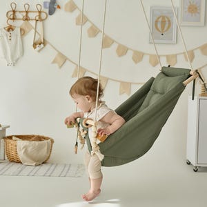 Op de afbeelding: Een olijfgroene babyhangmat die met touwen aan het plafond is opgehangen. Een baby zit in de hangmat en speelt met een houten speeltje. De kamer is ingericht met een neutrale inrichting, waaronder een witte kast, een kleine tafel en een rieten mand.