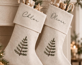May include: Two beige linen Christmas stockings featuring embroidered green pine trees and small white snowflakes. The cuffs are personalized with the names Chloe and Oliver in black script. Each stocking is filled with gifts, candy canes, and pine sprigs.