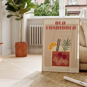 May include: A minimalist poster with the text "Old Fashioned" in a bold, red font. The poster features a stylized illustration of an old fashioned cocktail with orange slices, cherries, and ice cubes. The poster is framed in a gold frame and is hanging on a white wall.