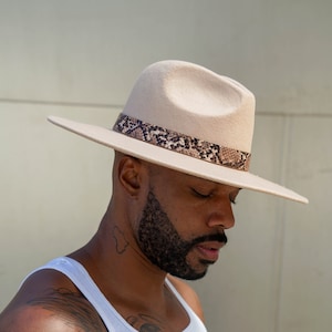 Może przedstawiać: Mężczyzna w białej koszulce bez rękawów z czarnym tatuażem na ramieniu i klatce piersiowej. Nosi beżowy kapelusz fedora z brązową wstążką w wzór skóry węża.