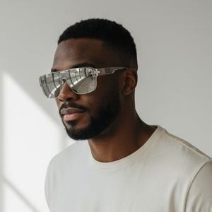 Puede incluir: Una persona con gafas de sol con montura plateada, una camiseta color crema y vaqueros azules. La persona est&aacute; de pie frente a una pared blanca con sombras.