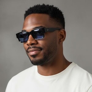 May include: Black square-framed sunglasses with blue lenses. The sunglasses are worn by a person wearing a cream-colored t-shirt. The background is a neutral gray.